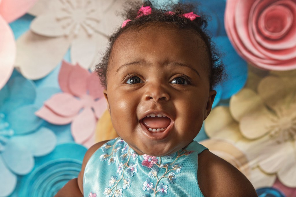 smiling baby portrait from first birthday cake smash with a girl wearing a dress with blue and pink flowers in front of a flowered backdrop in Prince Frederick Maryland
