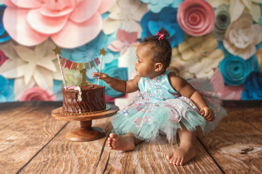 first birthday cake smash girl wearing blue and pink floral dress in front of backdrop with paper flowers and reaching towards her chocolate cake with a 1 and a banner with her name
