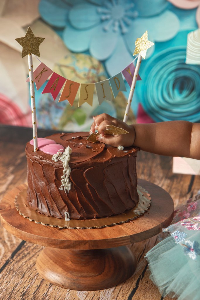 close-up of smash cake from first birthday photoshoot in Calvert County Maryland studio style with floral backdrop and baby reaching towards cake decorations