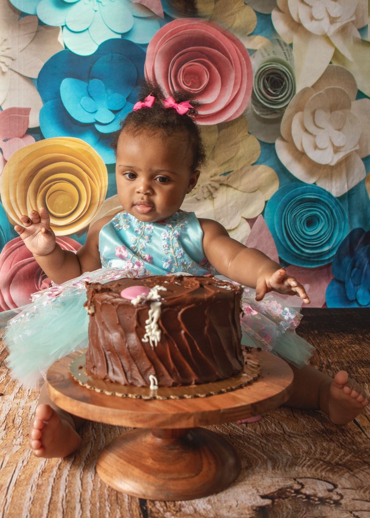 cake smash portrait in southern maryland of a baby girl sitting behind her chocolate cake while wearing a blue dress with pink and blue flowers